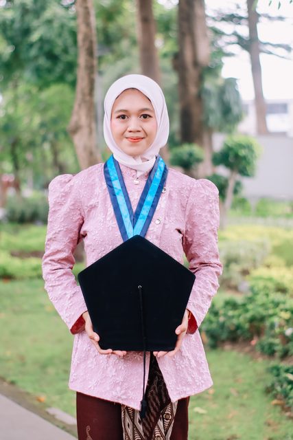 Wisudawati berkebaya merah muda memegang topi toga