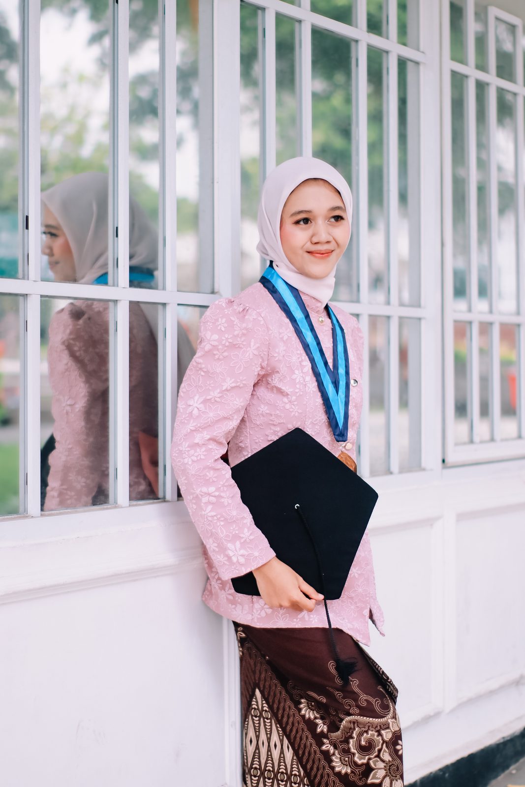 Wisudawati berkebaya merah muda di depan jendela putih