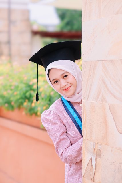 Wisudawati berkebaya merah muda mengintip dari balik tembok